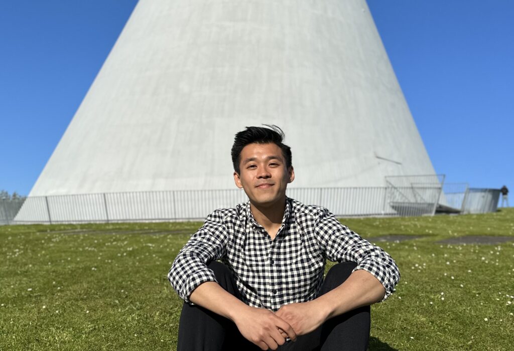 Henry is smiling while sitting on grass in front of a conically-shaped concrete structure. The sky is blue and clear in the background.