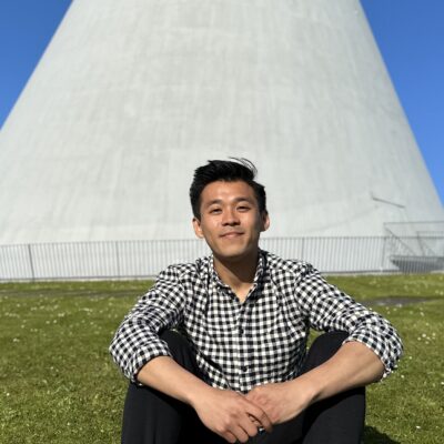 Henry is smiling while sitting on grass in front of a conically-shaped concrete structure. The sky is blue and clear in the background.