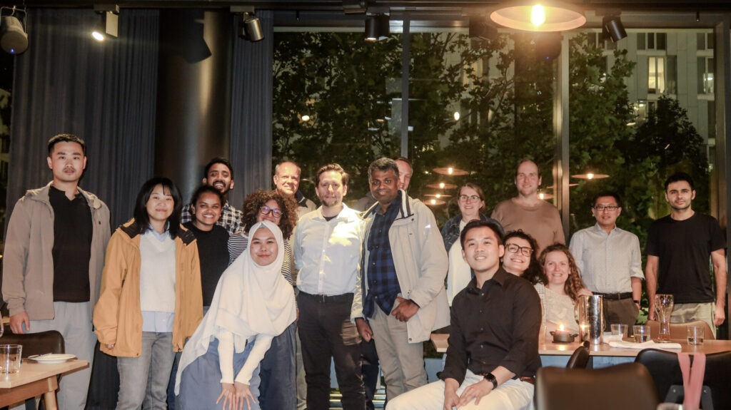 Photo of POSEIDON participants in a group smiling at the camera. The surroundings are dimly lit in a restaurant setting, with a couple of warm lights in the shot.