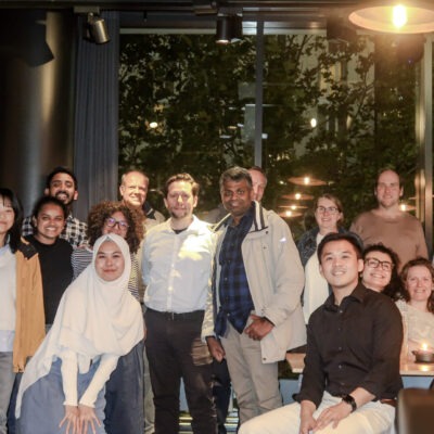 Photo of POSEIDON participants in a group smiling at the camera. The surroundings are dimly lit in a restaurant setting, with a couple of warm lights in the shot.