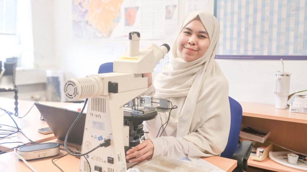 Photo of Hanifah sitting in front of a desk with a microscope; smiling at the camera.