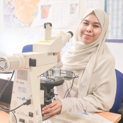 Photo of Hanifah sitting in front of a desk with a microscope; smiling at the camera.
