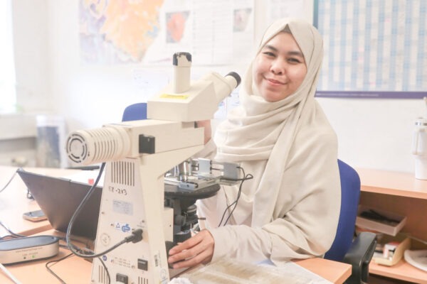 Photo of Hanifah sitting in front of a desk with a microscope; smiling at the camera.